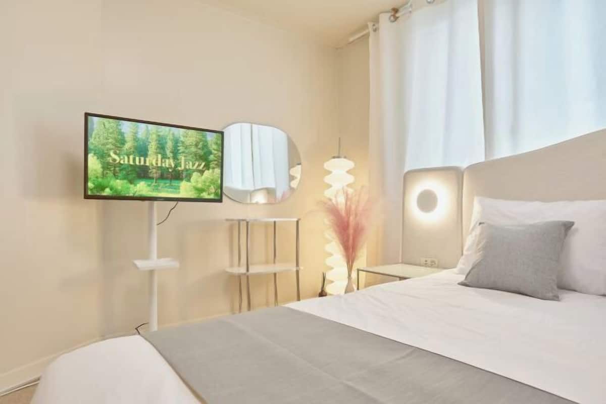Et soveværelse med seng, fjernsyn ophængt på væggen, et natbord og en lampe.