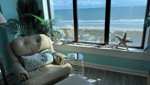Habitación con amplia ventana con vista a una playa, un sillón de cuero y una mesita con un adorno.