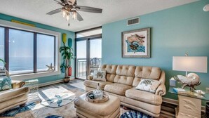 Un salón amplio con un gran ventanal, un ventilador de techo, un sofá color beige y un cuadro decorativo.