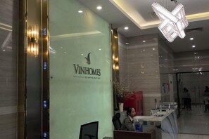 Un espace d’accueil doté d’un mur en verre sur lequel est affiché le logo « VINHOMES », d’un comptoir de réception et d’un lustre moderne.