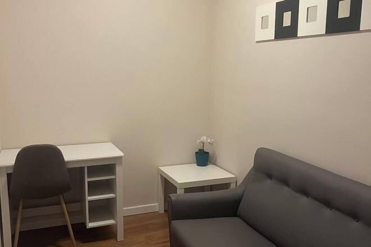 A corner room with a white desk, a grey sofa, a small white table with a blue vase, and a wall-mounted shelf with three black and white objects.