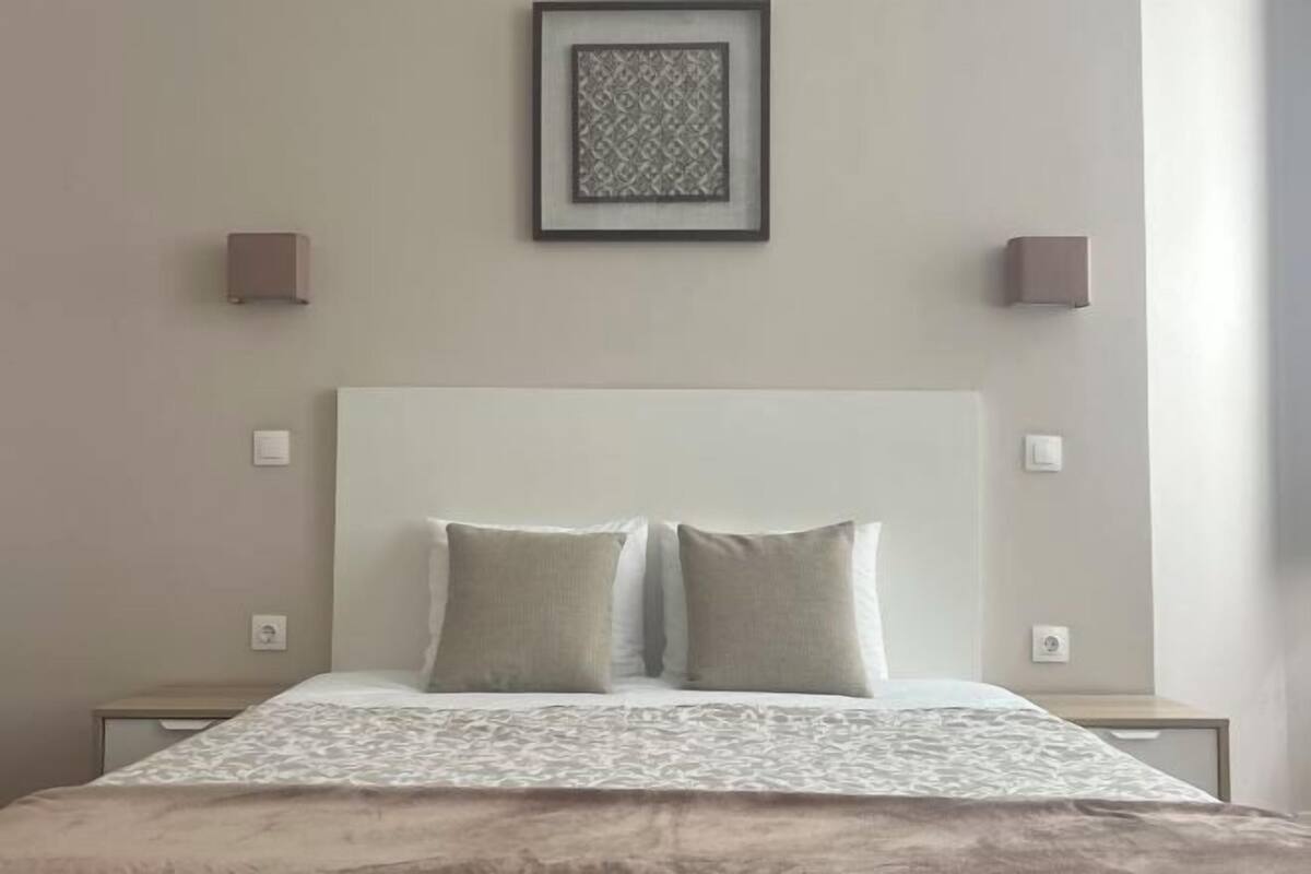 A neatly arranged bedroom with a large bed, two bedside tables, a framed artwork above the bed, and wall-mounted lights.