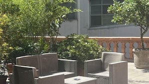 Des meubles de jardin en osier sont disposés sur une terrasse, agrémentée de plantes en pot, avec un bâtiment en arrière-plan.