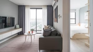 A modern hotel room with a sofa, a TV, a small table, and a balcony with curtains. - Luxury Apartment in Vinhomes D'Capitale by David Home (Hanoi)