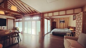 A spacious room with a wooden floor, a bed with a blue blanket, a sofa, a dining table with chairs, and a large window with sliding doors. - Traditional House Hotel Andong Villa (Andong-si)