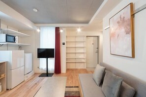 A modern living room with a sofa, a flat-screen TV, and a kitchen area with white cabinets and a microwave. - Apiche Oji (Tokyo)