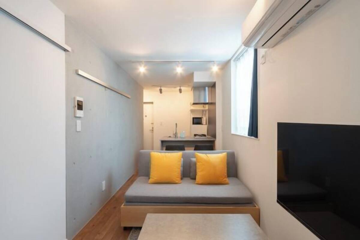 A narrow room with a sofa and cushions, a small table, and a kitchenette in the background.