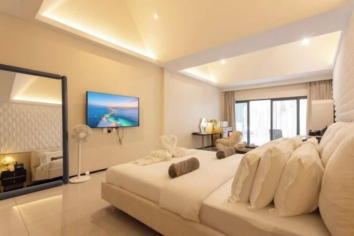 A modern hotel room with a large bed, a flat-screen TV, a desk with a lamp, and a large window.