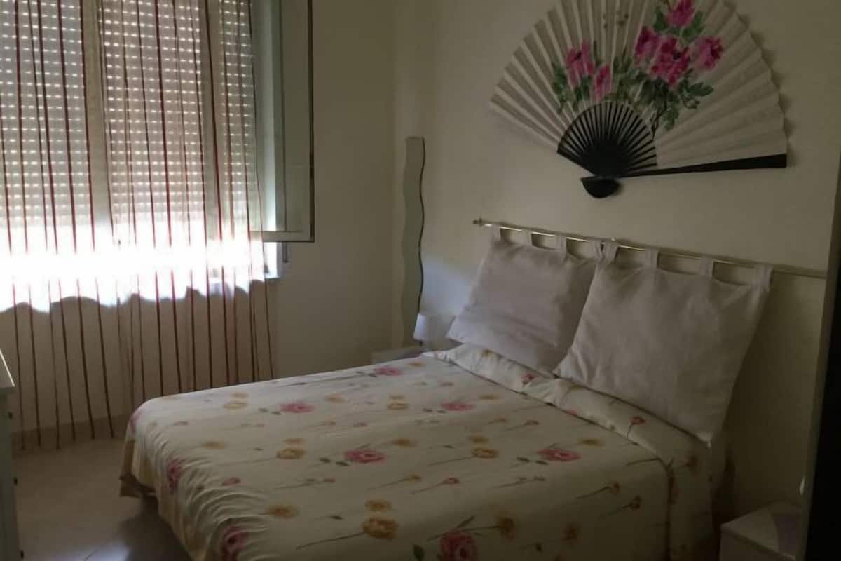 A bedroom with a bed, a window with blinds, and a wall-mounted fan with a floral design.