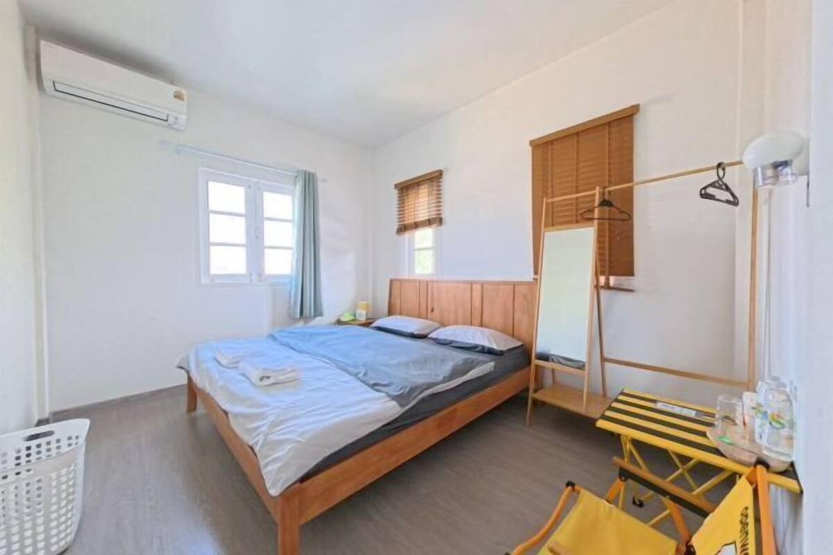 A bedroom with a bed, a wooden headboard, a window with blinds, a hanging light fixture, and a small table with a chair.