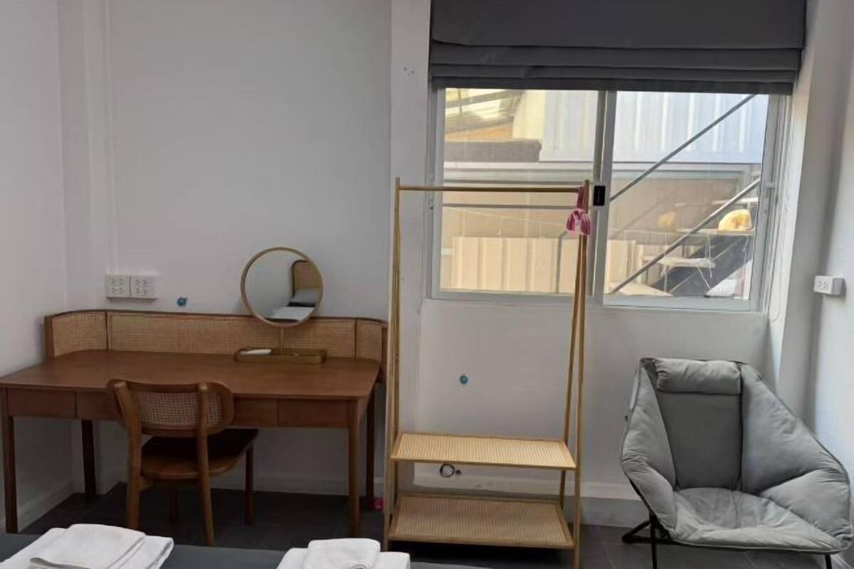 A room with a wooden desk, a chair, a bed, a window with a view, and a standing shelf.