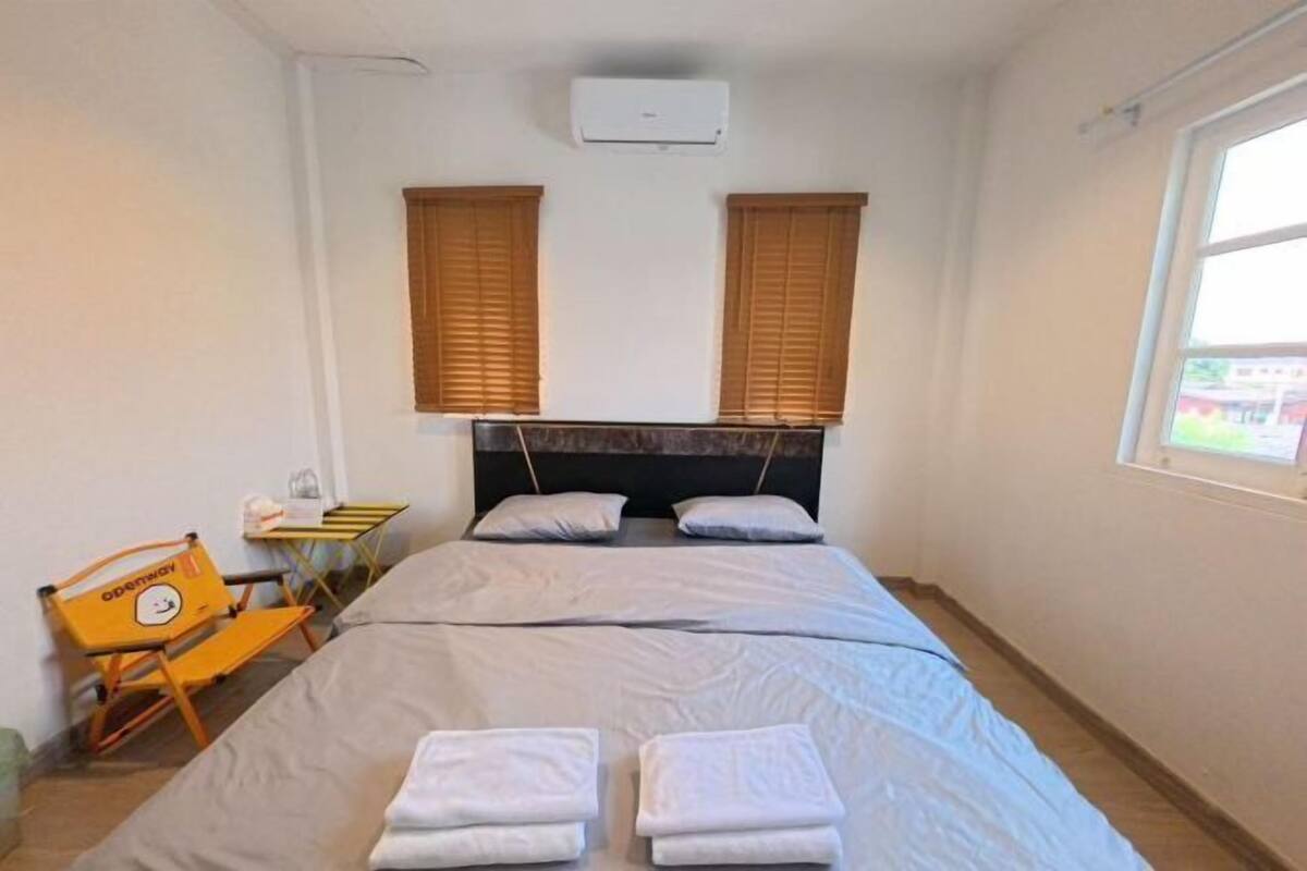 A hotel room with a bed, two pillows, a headboard, an air conditioning unit, a window with blinds, a chair, and a table with a towel.