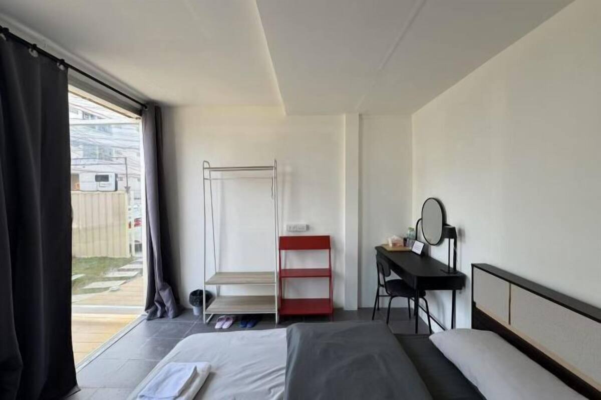 A hotel room with a bed, a desk with a chair, a mirror, a red shelf, and a window with curtains.