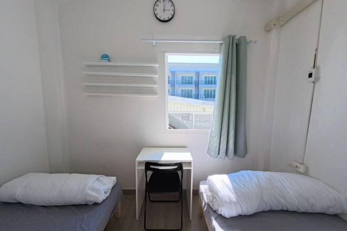 A small, clean room with two single beds, a desk, a chair, a clock, and a window with curtains.