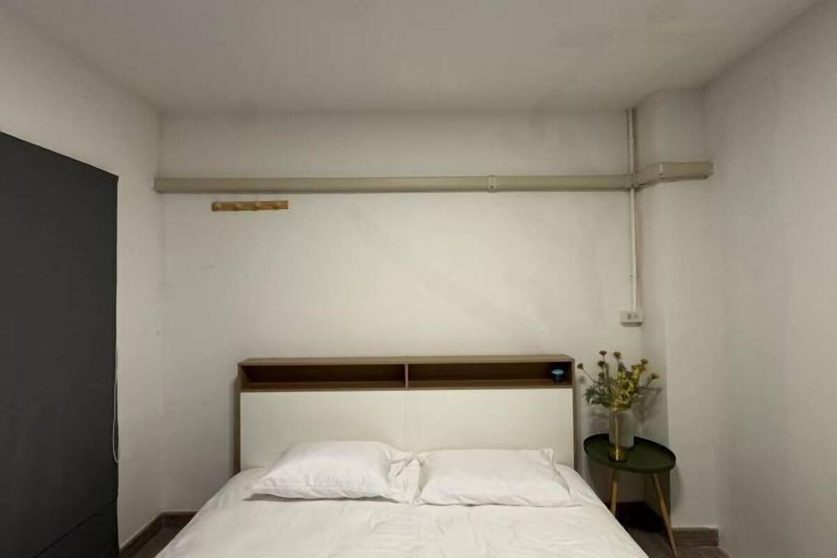 A simple, clean room with a bed, a small table with a vase, and a wooden headboard.