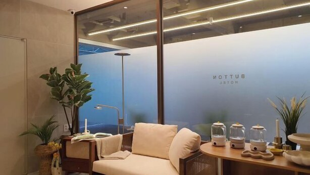 Een moderne hotelkamer met een bank, een bijzettafeltje met decoratieve objecten, een groot raam met uitzicht, en een muur met de naam van het hotel.