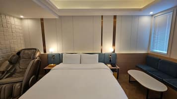 A modern hotel room with a large bed, massage chair, seating area, and a view of the city.