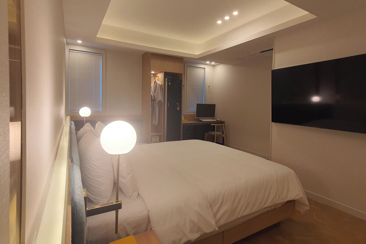 Un dormitorio con cama, lámpara de noche, escritorio con computadora, televisor montado en la pared y armario.
