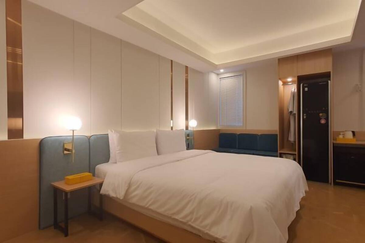 A hotel room with a large bed, bedside tables, a wardrobe, and a small desk with a lamp.
