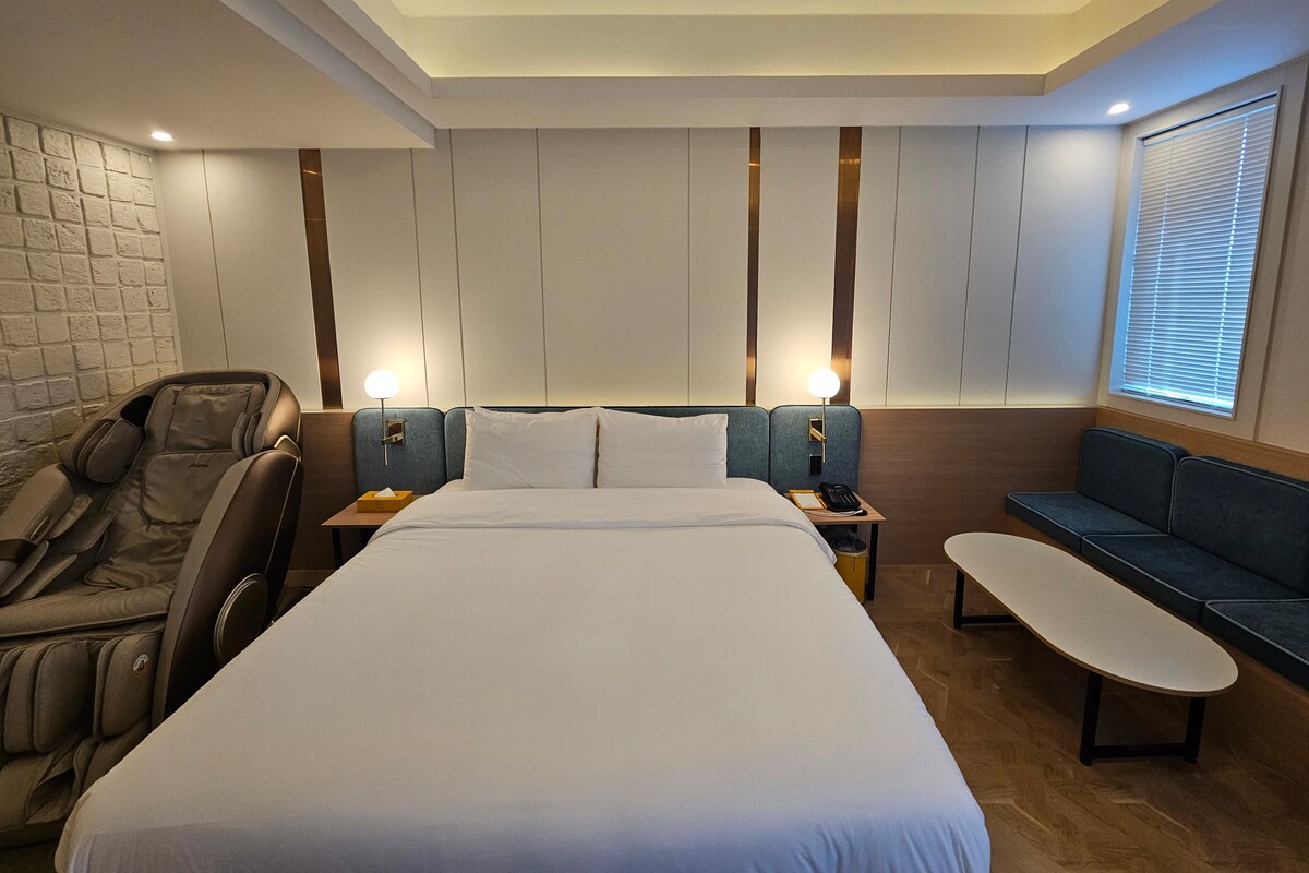 Una habitación de hotel con una cama grande, un sillón de masajes, una mesita de noche con lámpara, una pequeña mesa y un sofá.