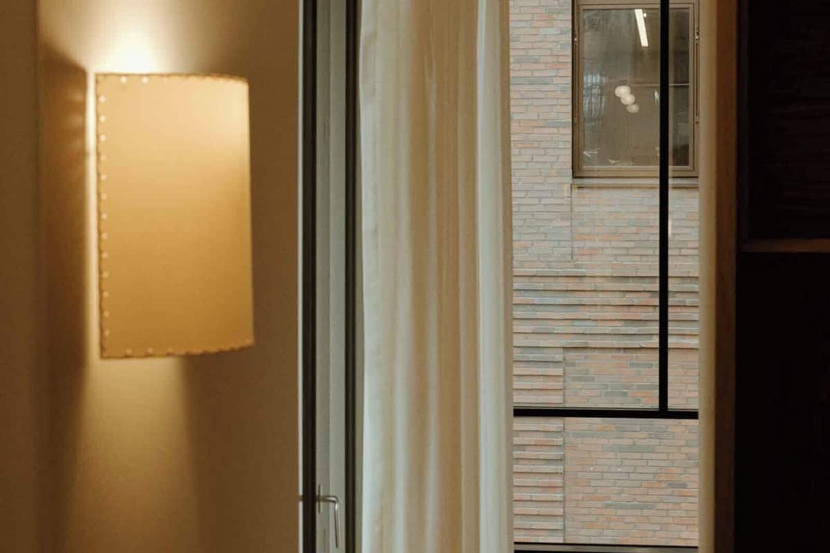 Un pasillo estrecho con una lámpara de pared encendida, un ventanal con persianas y una pared de ladrillo.