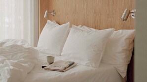 Ein ordentlich gemachtes Bett mit weißen Leinen und Kissen, ein kleiner Tisch mit Tasse und Buch sowie Holzvertäfelung an der Wand.