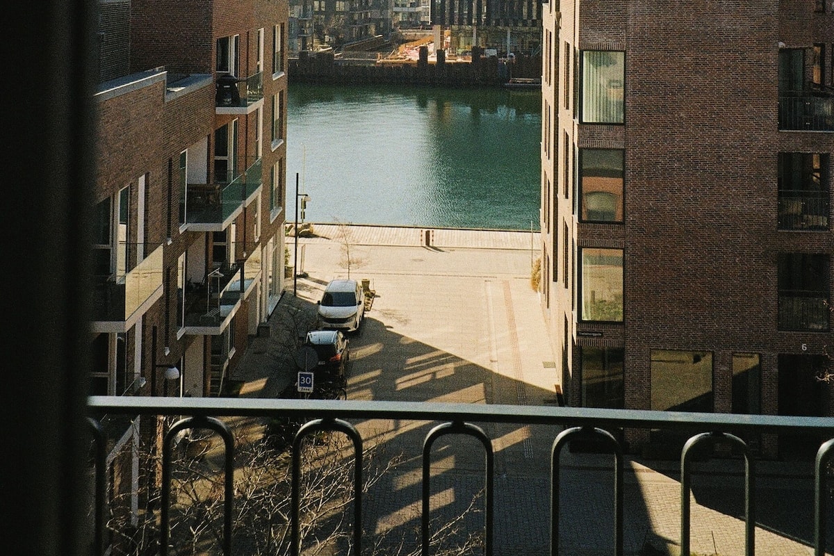 Vista de una zona urbana a orillas del río, con edificios modernos, cielo despejado y una barandilla metálica al frente.