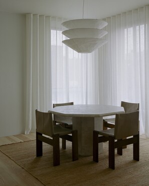 Un comedor con una mesa redonda, seis sillas, una lámpara colgante y cortinas blancas traslúcidas.