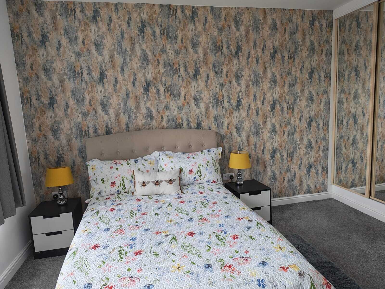 Un dormitorio con colcha estampada, dos mesitas de noche con lámparas, un cabecero y un papel pintado con diseño.