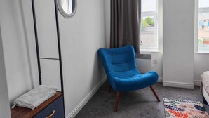 Un dormitorio con una silla azul, estante de madera, espejo, ventana con cortinas y una alfombra colorida.