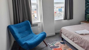 Un dormitorio con una silla azul, una cama con colcha estampada y vistas a la calle a través de dos ventanas con cortinas grises.