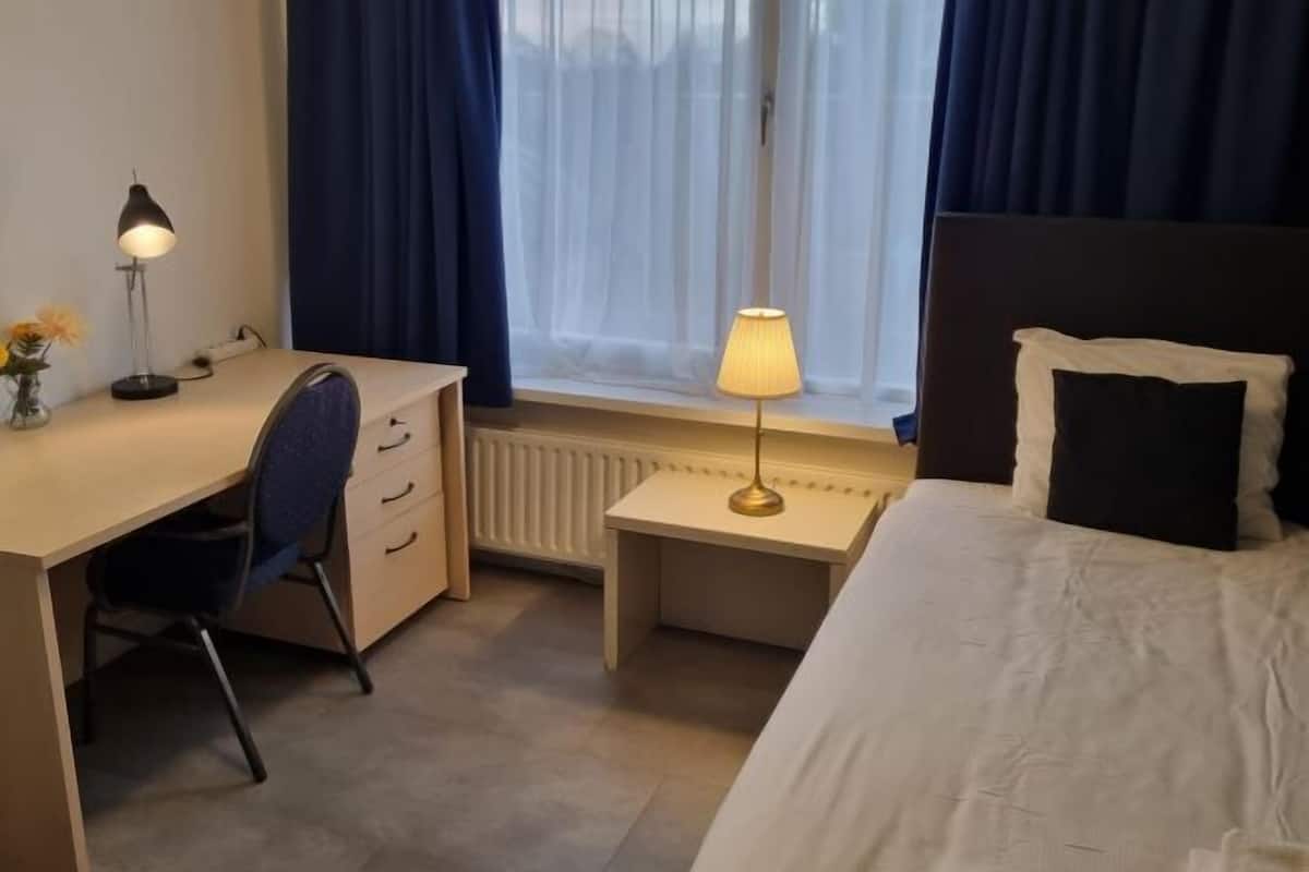 Une chambre d’hôtel avec un lit, un bureau, une chaise, une lampe et une fenêtre avec des rideaux.
