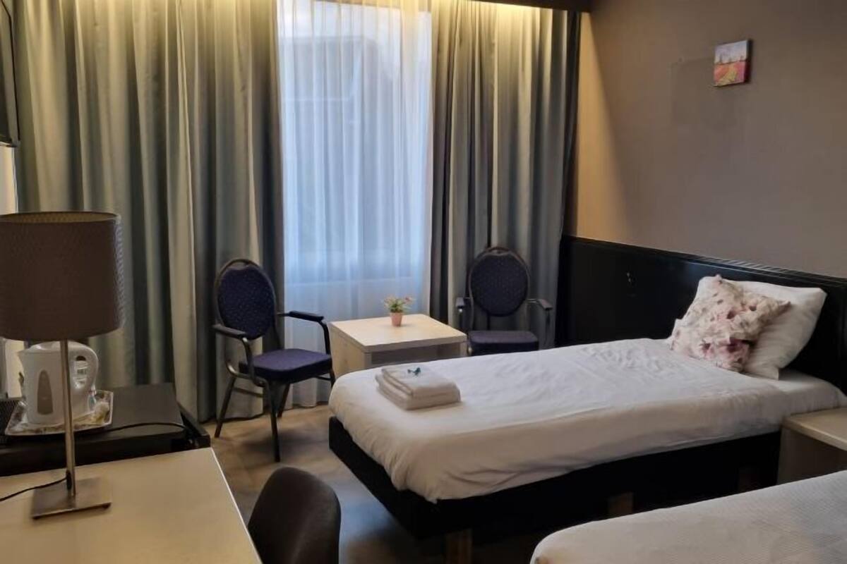 Une chambre d’hôtel avec deux lits, un bureau avec une lampe, une chaise, une petite table avec une plante et une fenêtre avec des rideaux.