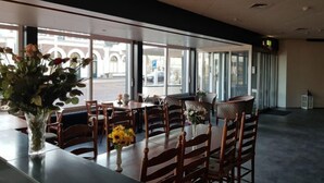 Um restaurante com mesas e cadeiras de madeira, janelas amplas e um vaso com flores.
