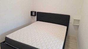 A small, single-bed room with a white mattress and a black headboard. There is a small shelf unit on the wall to the right of the bed. - Kibou Apartment (Garbagnate Milanese)