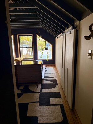 A narrow hallway with a wooden floor, a black and white rug, a wooden bench, and a window with a view of trees. - Raven Cove / Modern Lakefront Retreat (Findley Lake)