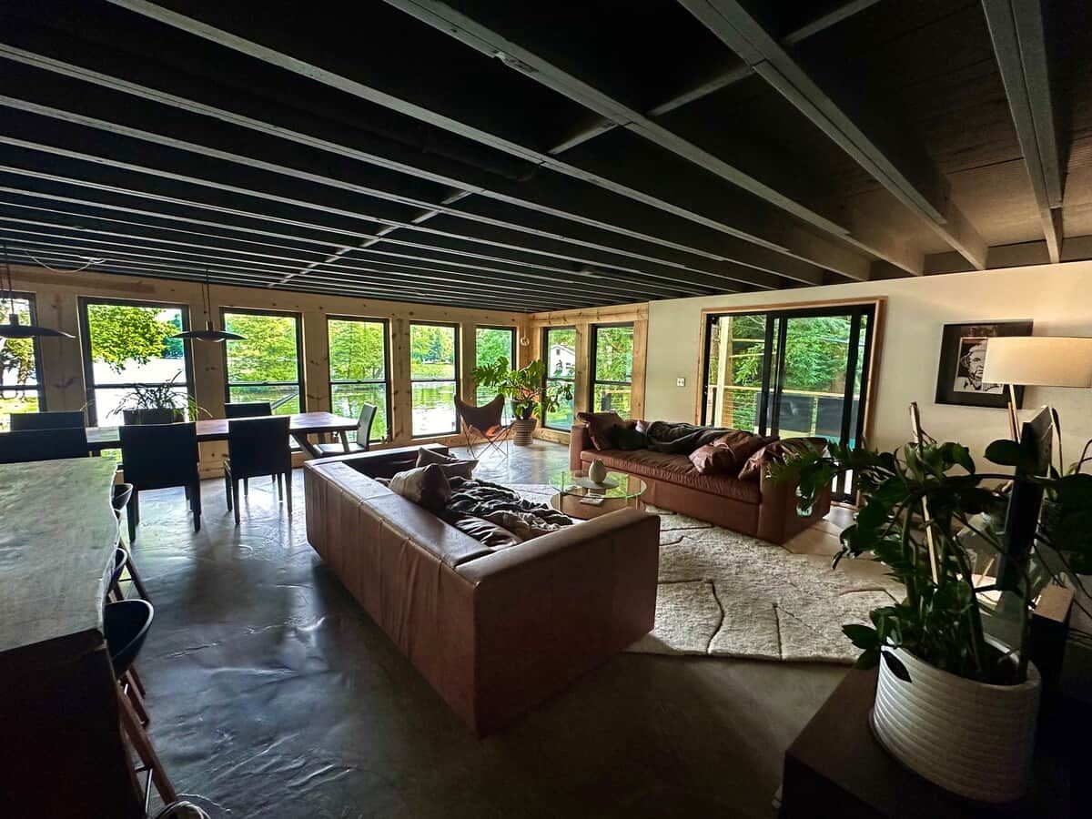 A spacious living area with a high ceiling, large windows, and a mix of seating arrangements including a sofa, a couch, and a dining table with chairs.