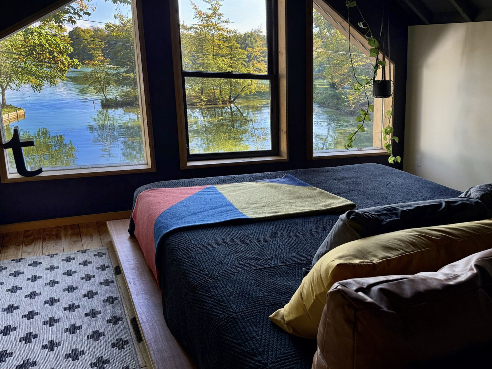 Un dormitorio con un ventanal grande que da a un lago, una cama con colcha y suelo de madera.