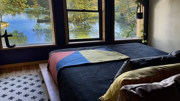 Un dormitorio con un ventanal grande que da a un lago, una cama con colcha y suelo de madera.