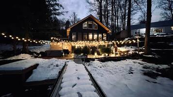 Una cabaña de madera con porche cubierto, rodeada de nieve, y luces colgantes.