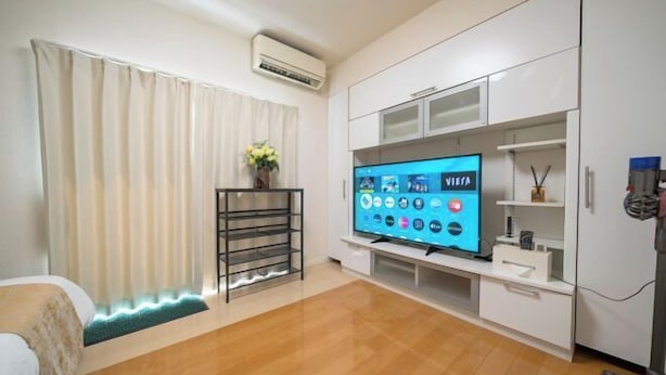 Una sala de estar moderna con televisor de pantalla plana, muebles blancos, piso de madera y una ventana con cortinas.