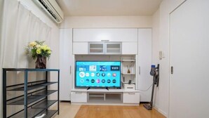 Una habitación con una televisión de pantalla plana montada sobre un mueble blanco, una estantería metálica a la izquierda y una aspiradora a la derecha.