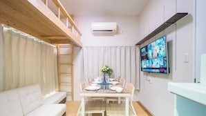 Un espacio habitable compacto con una mesa de comedor lista para seis personas, un sofá, un televisor montado en la pared y un altilo en madera sobre el sofá.