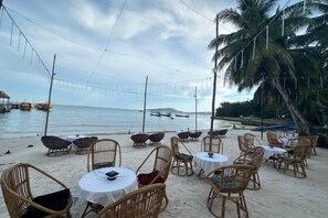 Ein Essbereich am Strand mit Korbstühlen und Tischen, mit Blick auf das Meer.