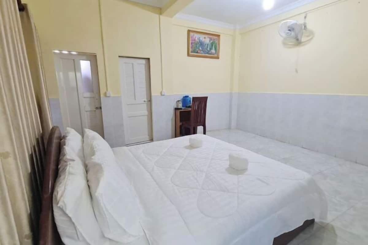 A hotel room with a bed, a door, a small table with a water dispenser, and a wall-mounted fan.