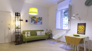 A modern living room with a green sofa, a dining table with a bowl, a white chair, a yellow lamp, and a window with a view of trees. - Fico d'India by Wonderful Italy (Siracusa)