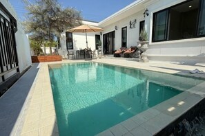Ein Schwimmbecken mit Fliesenrand, eine Terrasse mit Gartenmöbeln und ein Gebäude mit großen Fenstern.