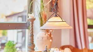 A hanging lamp with a white shade, a vase with flowers, and a window with a view of buildings. - Mercure Hotel Bad Duerkheim An Den Salinen (Bad Durkheim)