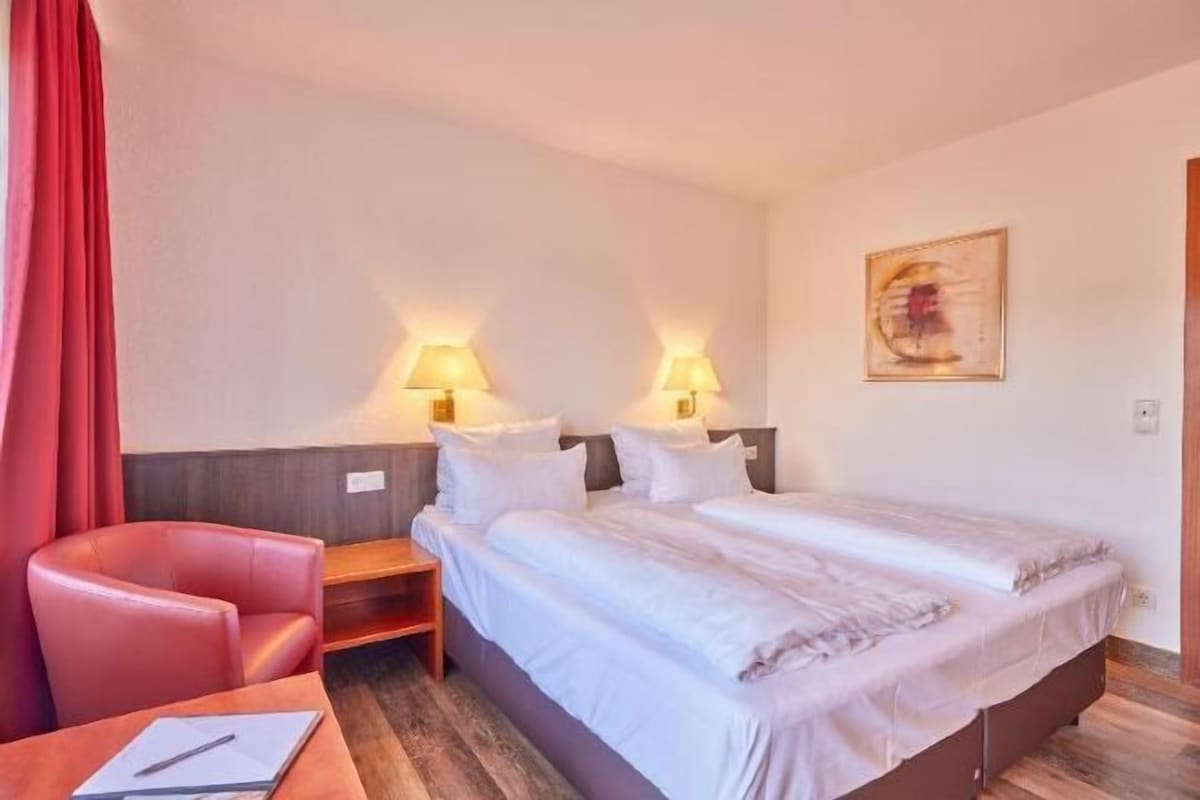 Ein Hotelzimmer mit einem Bett, zwei Nachttischlampen, einem Sessel, einem Schreibtisch und einem Bild an der Wand.