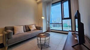 Un salon moderne avec un canapé, une table basse ronde, un téléviseur et une grande fenêtre donnant sur la ville.